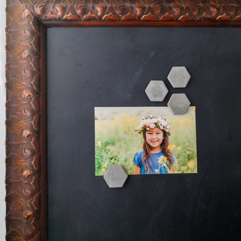 4 hexagon shaped concrete magnets holding a photo of a young girl in a field of flowers onto a magnetic chalkboard with a baroque style picture frame