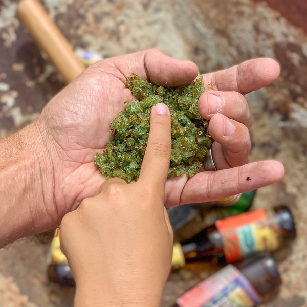 Rough manly hand with fingers forming rock sign language symbol holding green and brown pulverized beer bottle glass and a kids hand reaching in to touch the recycled concrete aggregate to be with bottles in the background